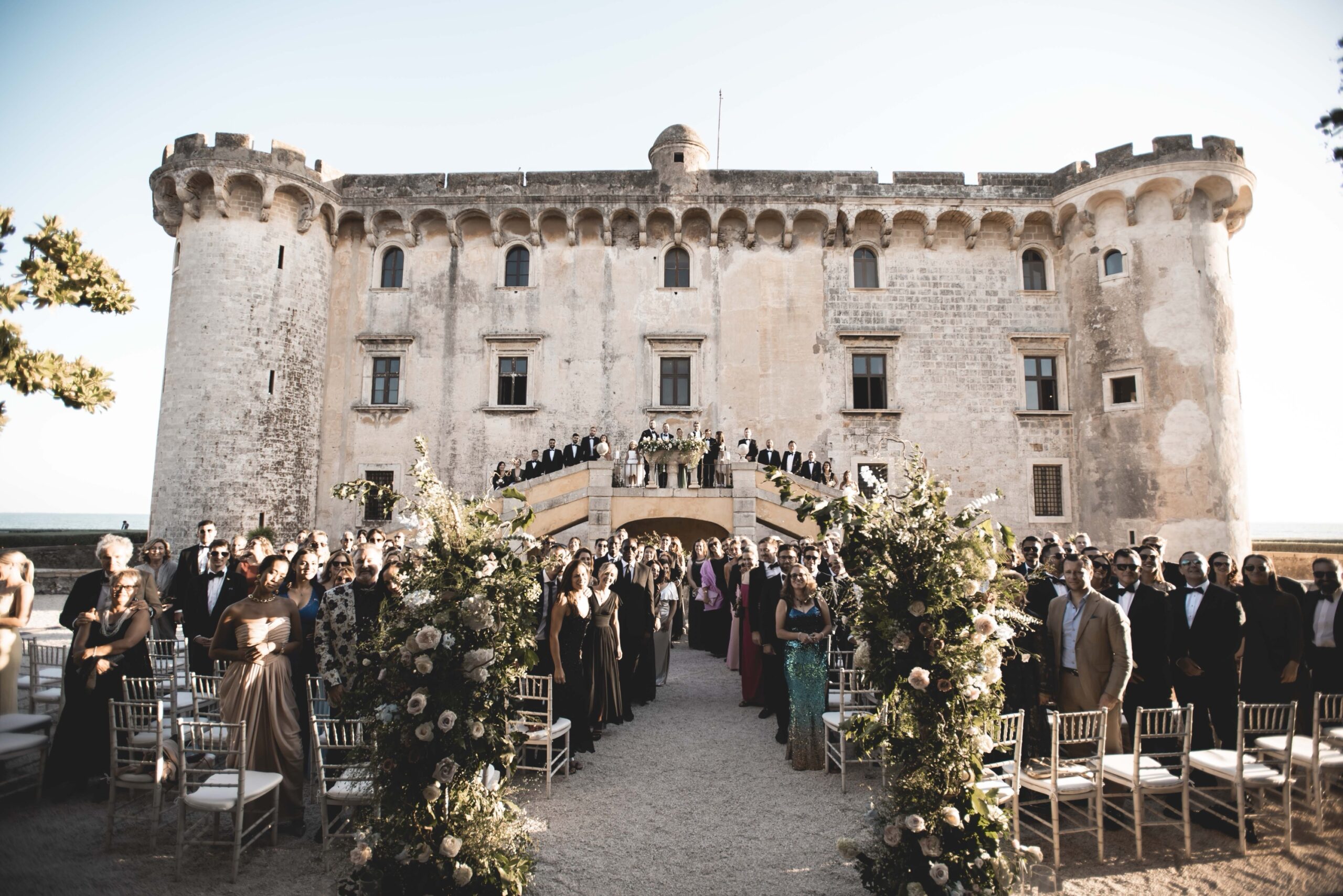 Guests standing for an outdoor ceremony in front of the Castle of Palo, the aisle framed by tall floral arrangements, Diamante Your Wedding in Italy