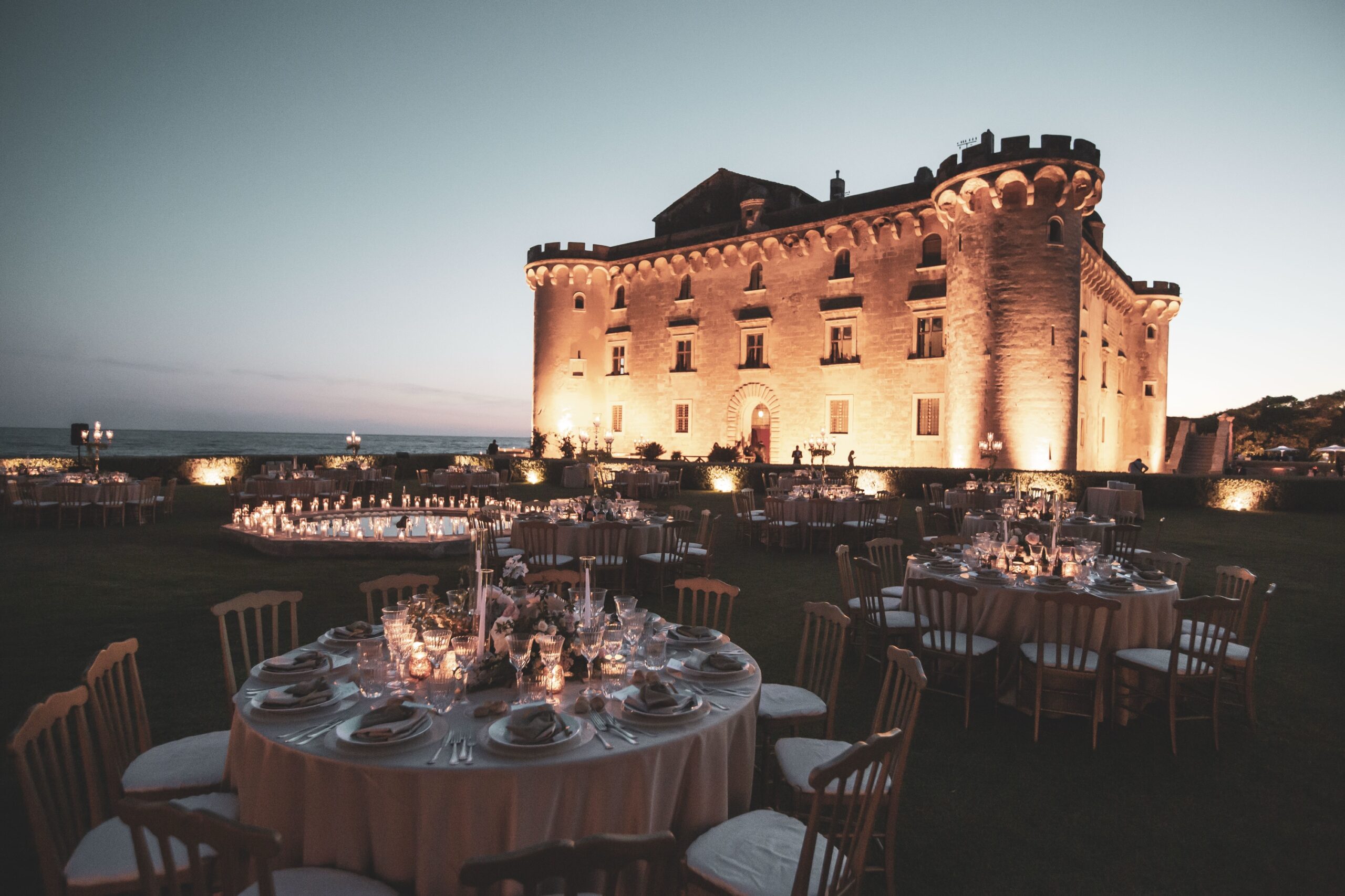 Round dinner tables set on the castle lawn at Palo Laziale as the lights come on at dusk, Diamante Your Wedding in Italy
