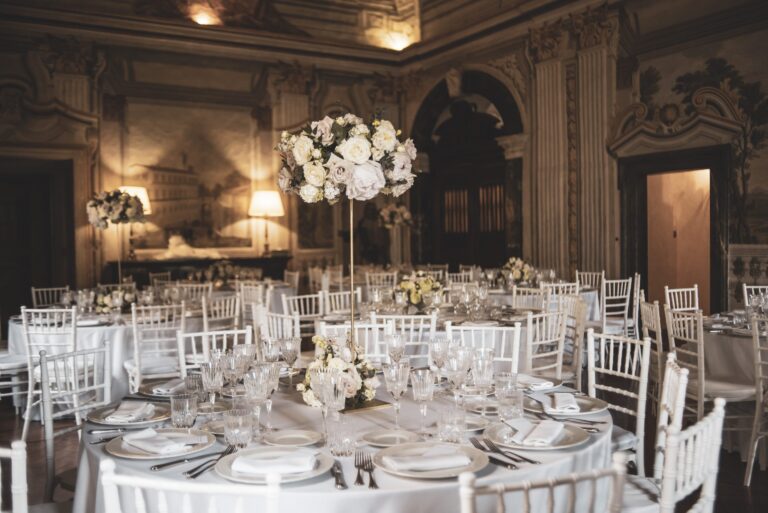 Round tables with tall white rose centrepieces in a frescoed hall, Diamante Your Wedding in Italy