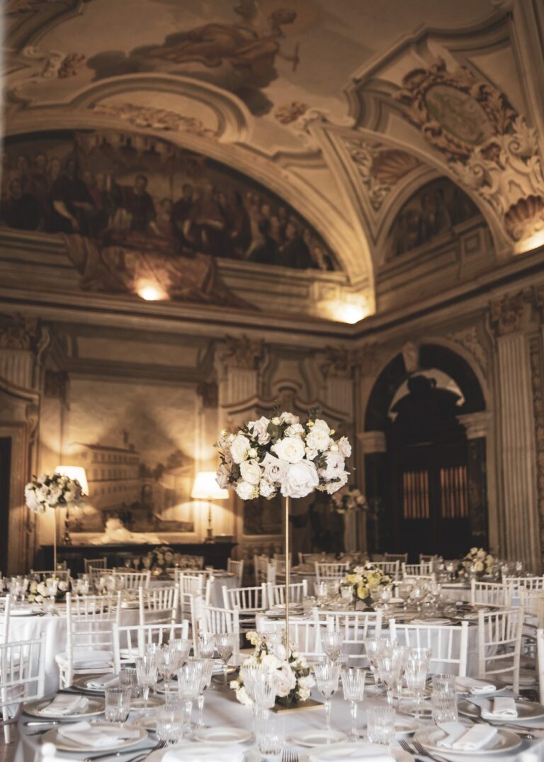 A painted vaulted hall set with white tables and raised rose centrepieces, Diamante Your Wedding in Italy