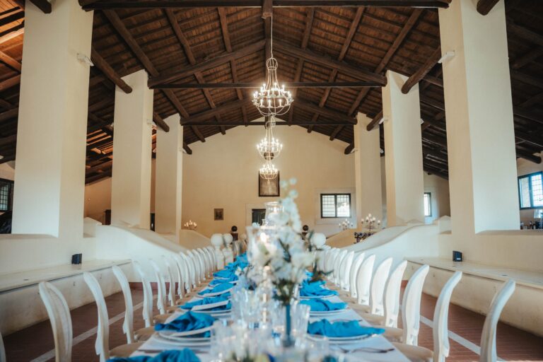 A long table with blue napkins and white flowers under the beamed roof of the event hall at Tenuta di Fiorano, Diamante Your Wedding in Italy