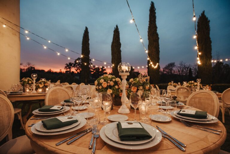 A round table with green napkins and peach roses under festoon lights and cypresses at sunset, Tenuta di Fiorano, Diamante Your Wedding in Italy