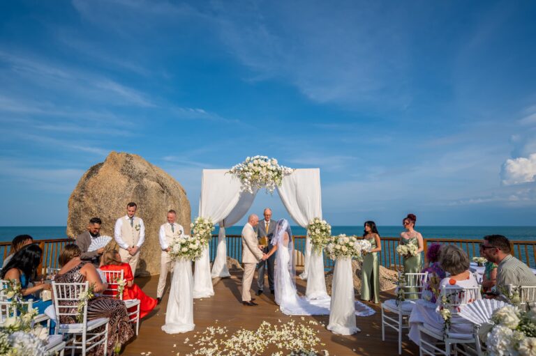 A seaside terrace ceremony with a white floral arch and rocks behind, photographed from above, Dimas Frolov Photography