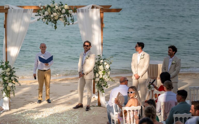 A beach ceremony beneath a wooden arch dressed with white flowers and fabric, Dimas Frolov Photography