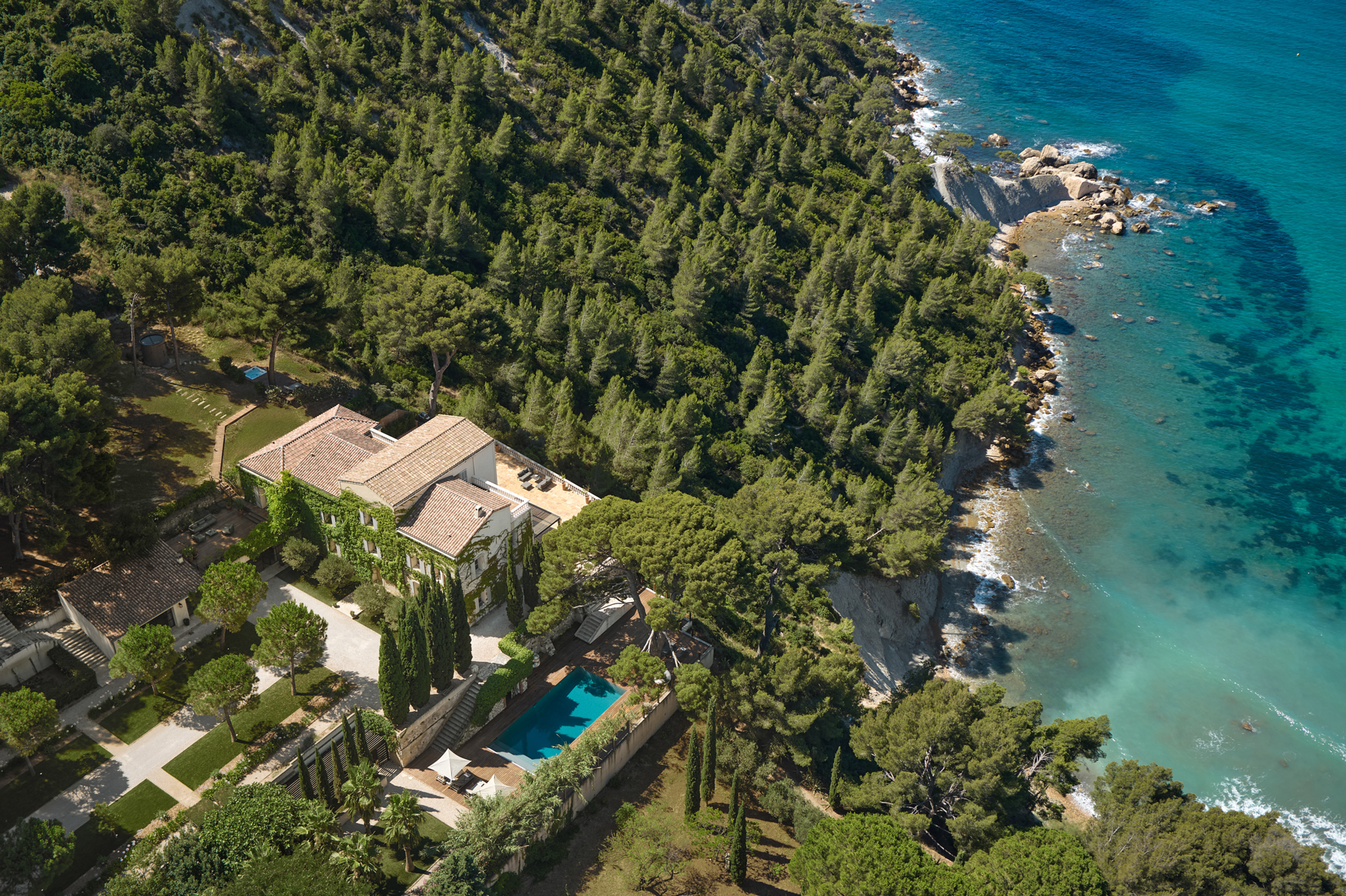 Aerial view of the villa among pine trees above the turquoise sea, with the pool below the house