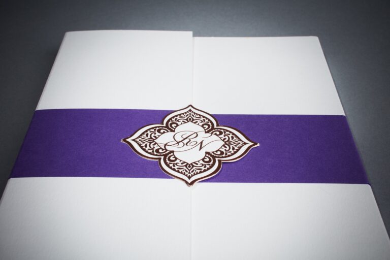 Cream ceremony invitation wrapped in a purple ribbon with a copper foil engraved mandala monogram, Downey Luxury Stationery