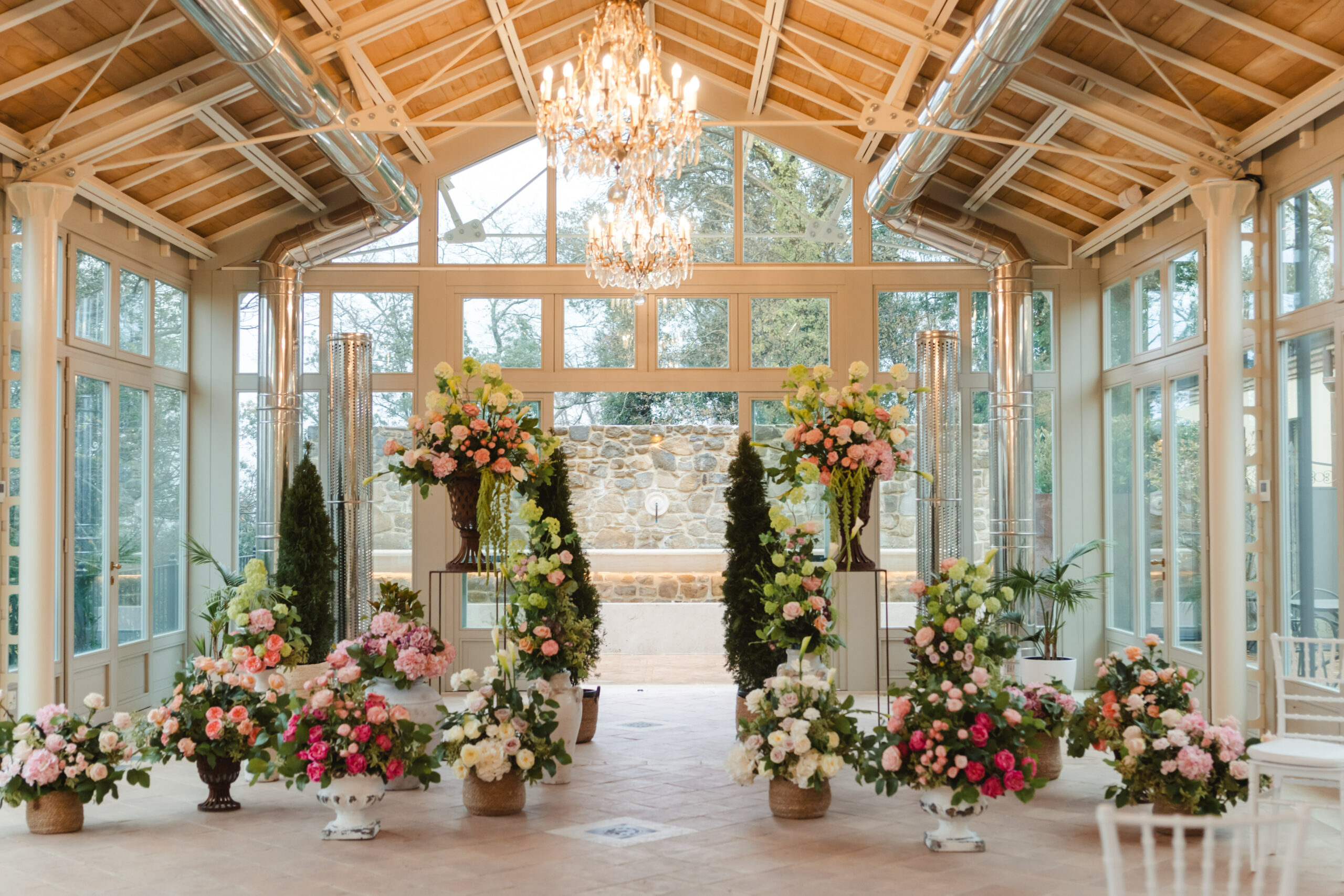 A glasshouse under a timber roof set for a ceremony: a crystal chandelier, urns of pink and coral flowers lining both sides of the aisle, Dream On Wedding Team