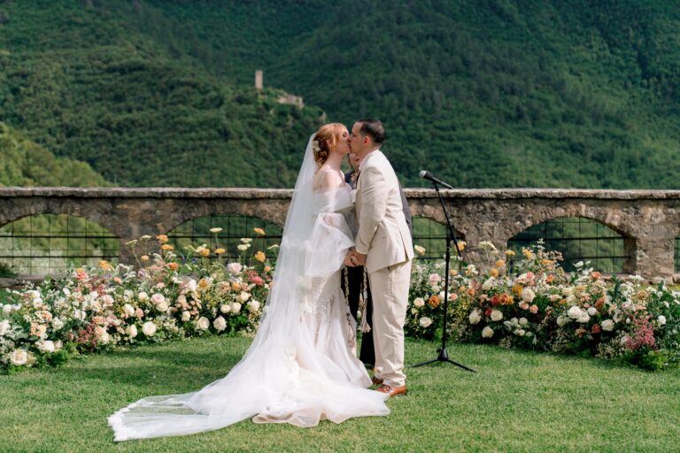 An outdoor ceremony on a lawn, a low border of peach, cream and pink flowers along an arcaded stone wall, a wooded hillside behind, Dream On Wedding Team