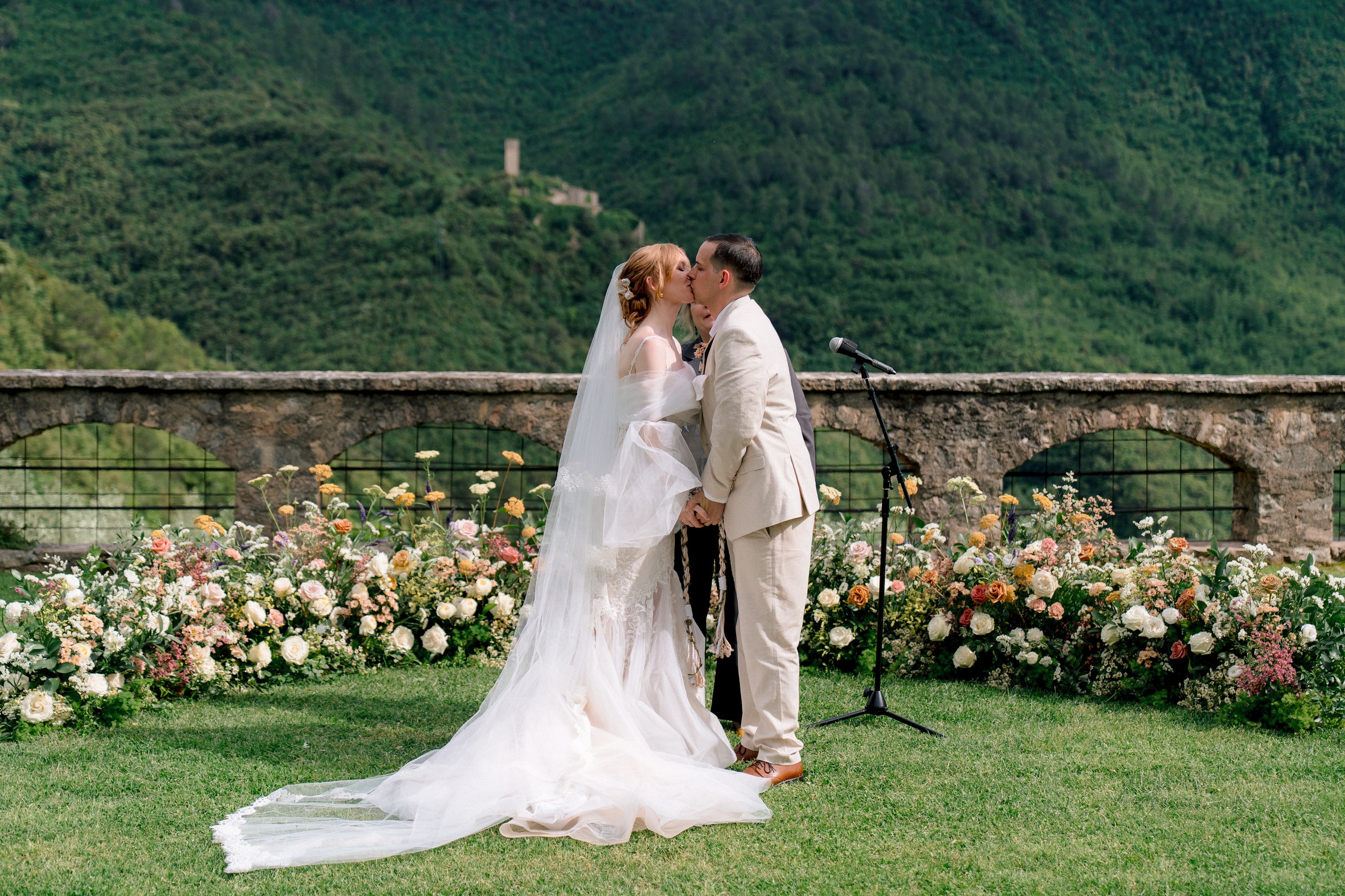 An outdoor ceremony on a lawn, a low border of peach, cream and pink flowers along an arcaded stone wall, a wooded hillside behind, Dream On Wedding Team