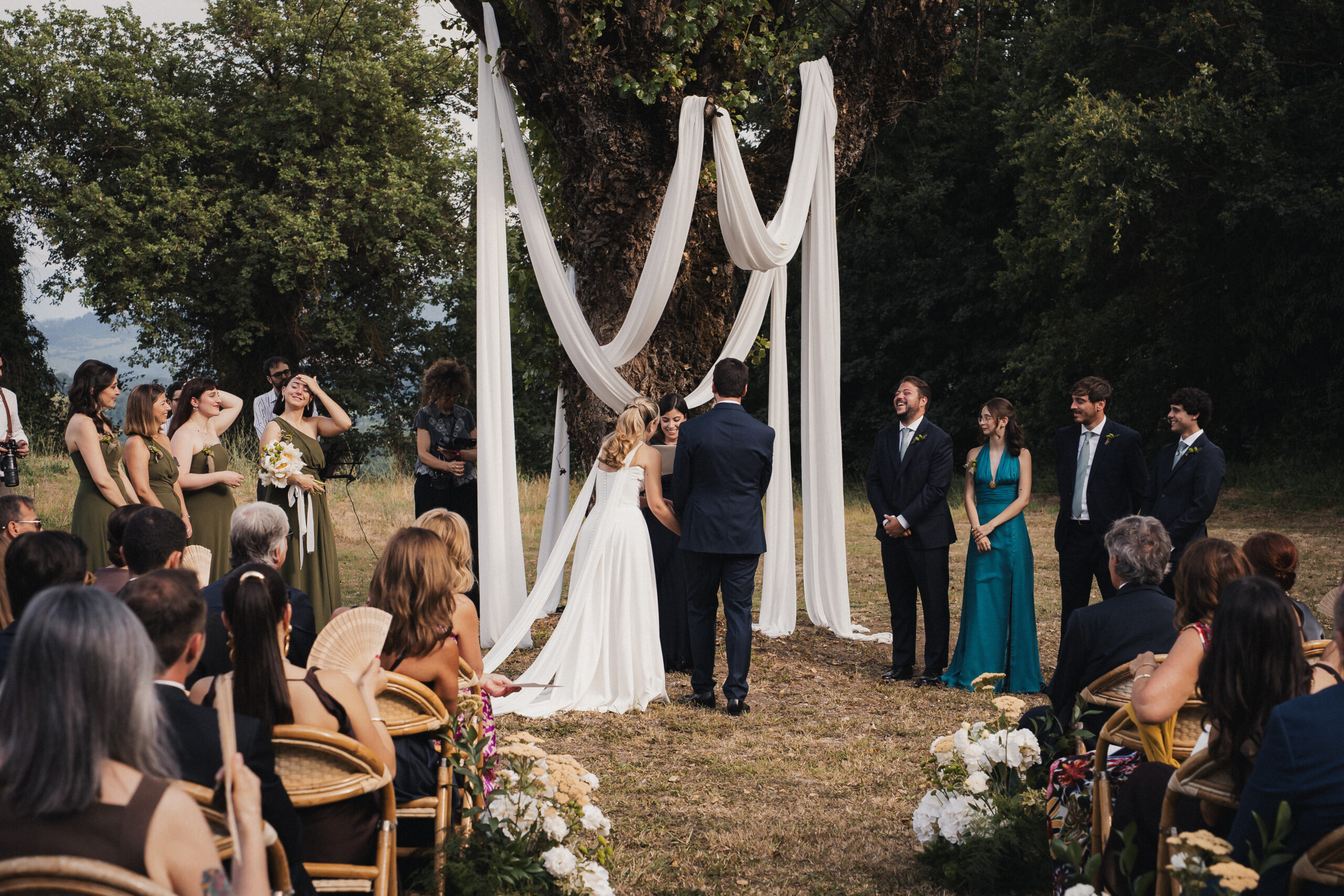 A ceremony beneath a large tree, a white draped frame behind the couple, guests seated on cane chairs, Dream On Wedding Team