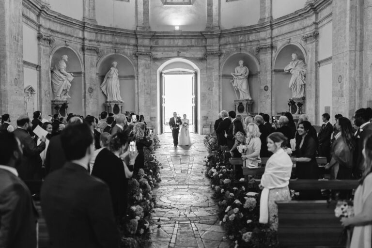 Black and white: a domed rotunda church with statues in the niches, flowers along the aisle and the bride at the open door, Dream On Wedding Team