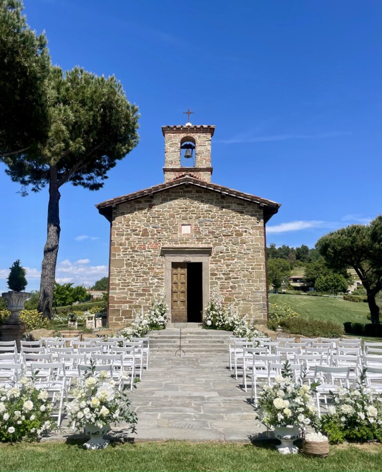 A small stone country chapel with a bell gable, white chairs and flowers set out on the lawn in front, umbrella pines either side, Dream On Wedding Team