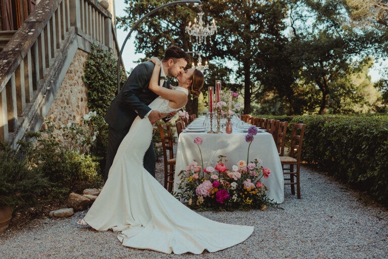 A table for two on a gravel terrace with tapers and a low garden arrangement, a chandelier hung in the trees above, Dream On Wedding Team