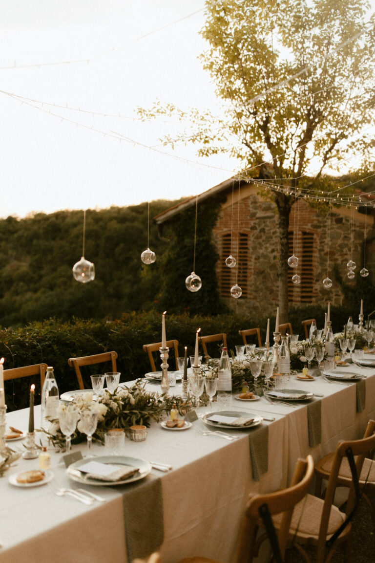 A long dinner table laid beneath a tree at sunset, glass baubles hanging from the branches, a stone building behind, Dream On Wedding Team