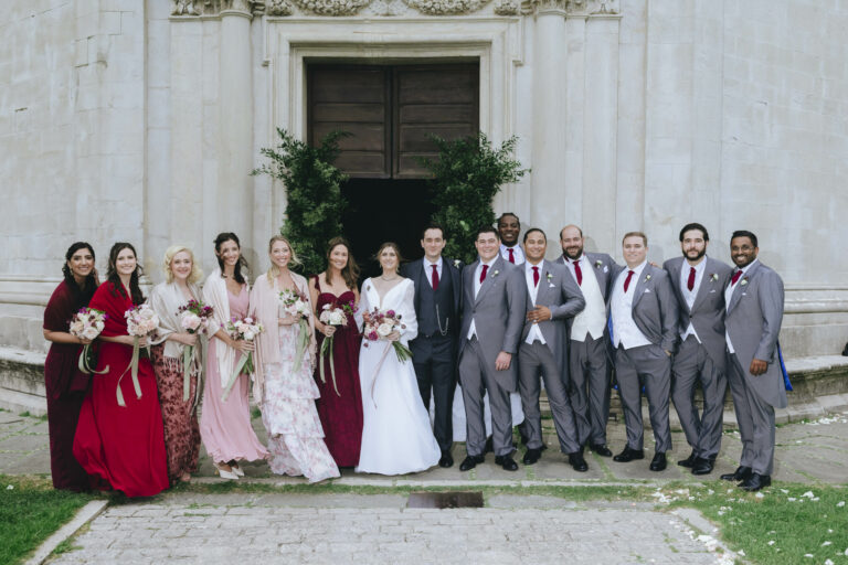 The wedding party in front of a pale stone church: bridesmaids in burgundy and blush, groomsmen in grey, Dream On Wedding Team