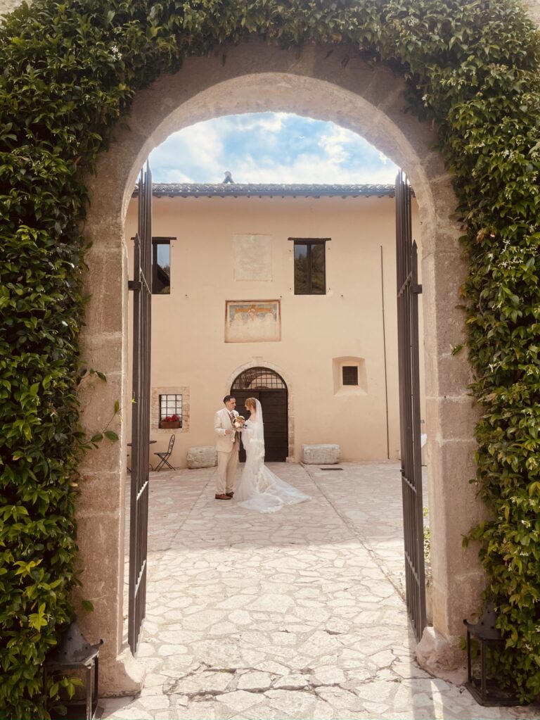 An ivy-hung stone archway opening onto a courtyard, the couple standing in the doorway of the house beyond, Dream On Wedding Team