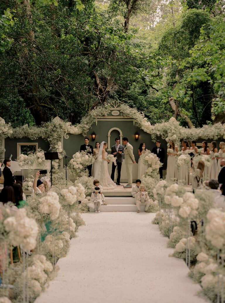 Couple walking up an aisle of white flowers through the garden ceremony, Dream Weddings Europe