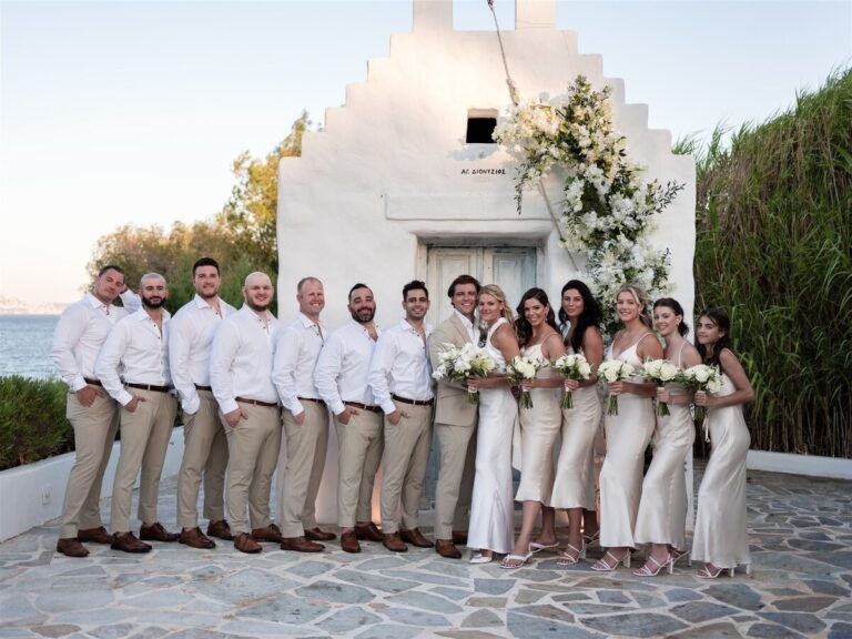 A wedding party lined up in front of a whitewashed chapel with a floral arch, Dream Weddings Mykonos