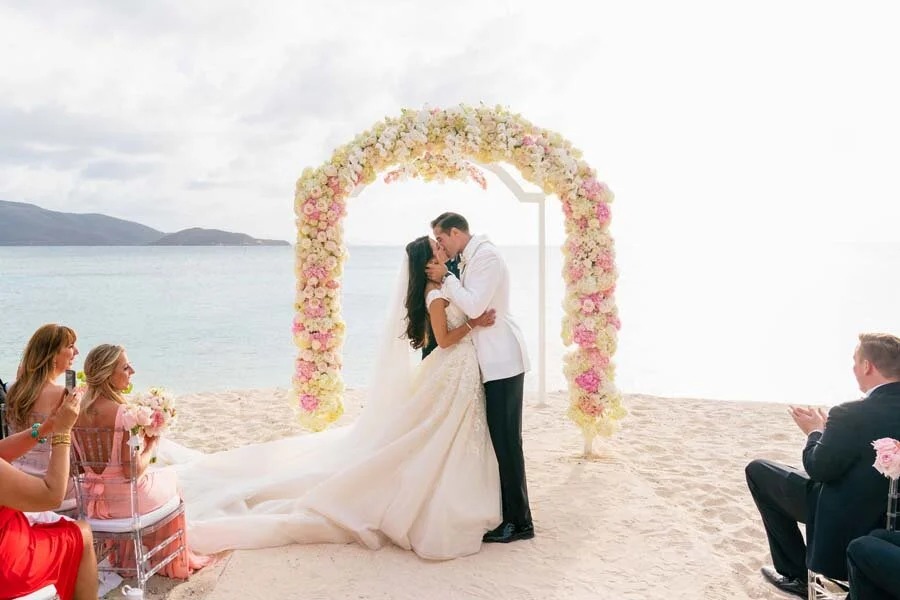 A couple kissing beneath a pastel floral arch on the sand, Necker Island