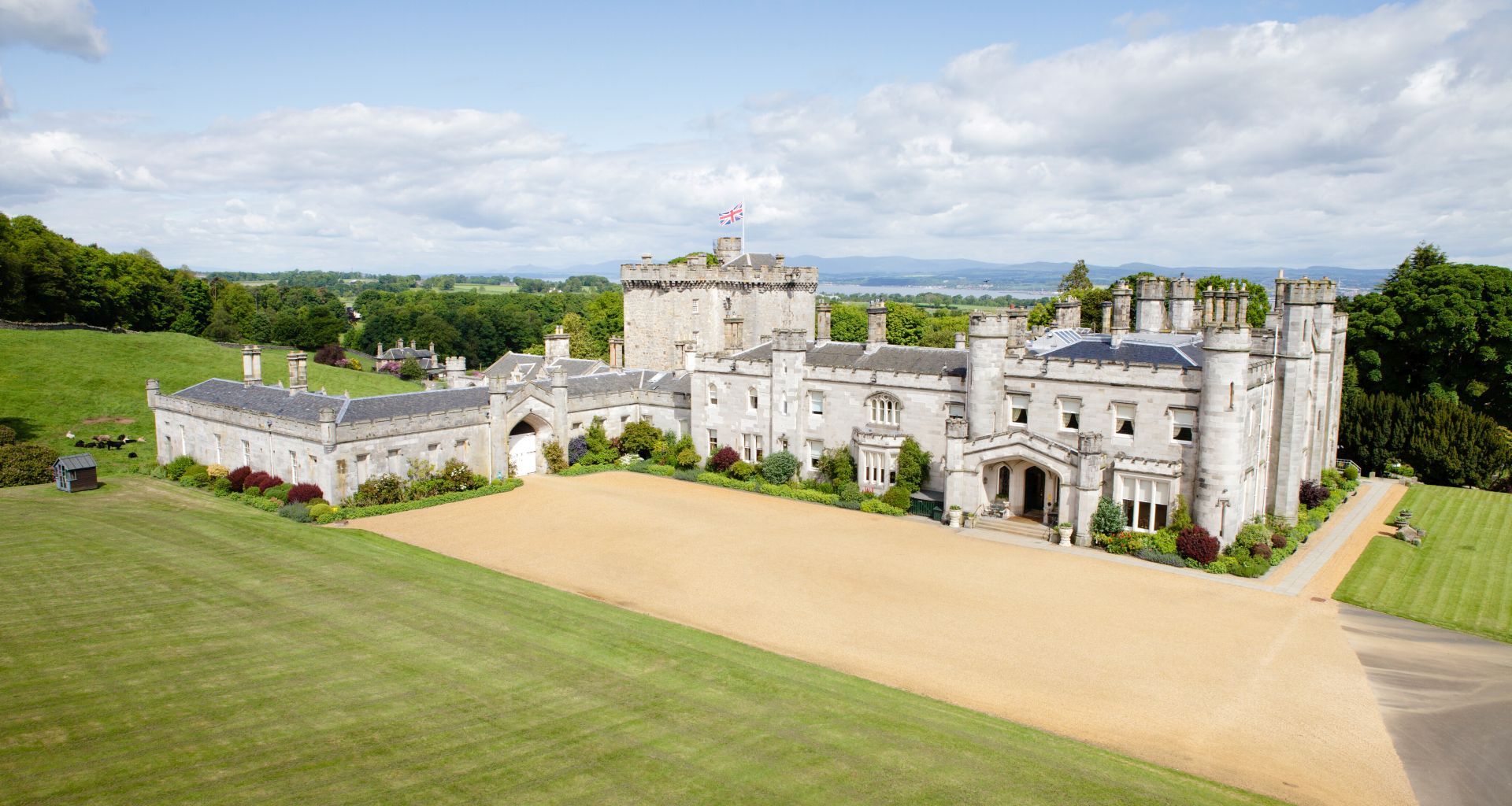 The castle from the air across the front lawn and gravel forecourt, with the Auld Keep behind and the Firth of Forth on the horizon, Dundas Castle