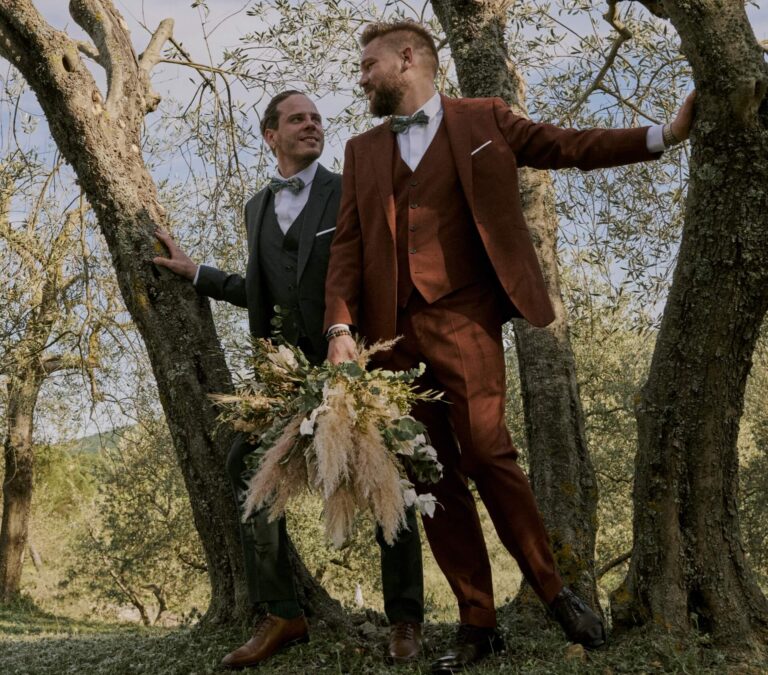 Two grooms in an olive grove in Tuscany, burgundy and dark suits, a dried-flower bouquet, Elisa Prati Wedding Italy