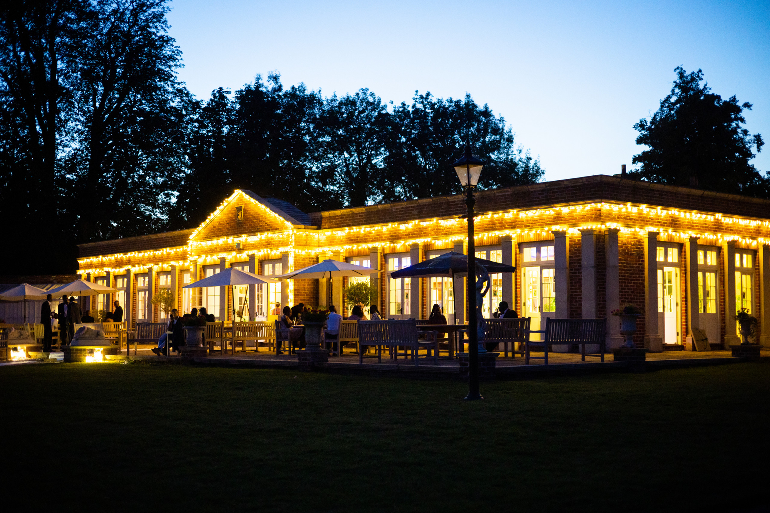 The Orangery lit with festoon lights at dusk, with guests on the terrace under parasols, Elmhay Park