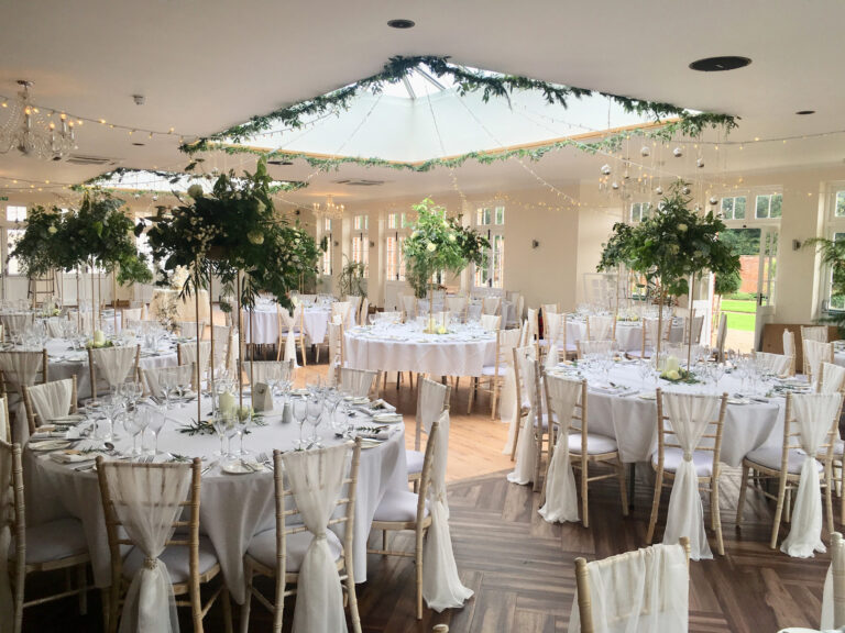 Round tables laid for a wedding breakfast in the Orangery, with tall green centrepieces under the roof lantern, Elmhay Park