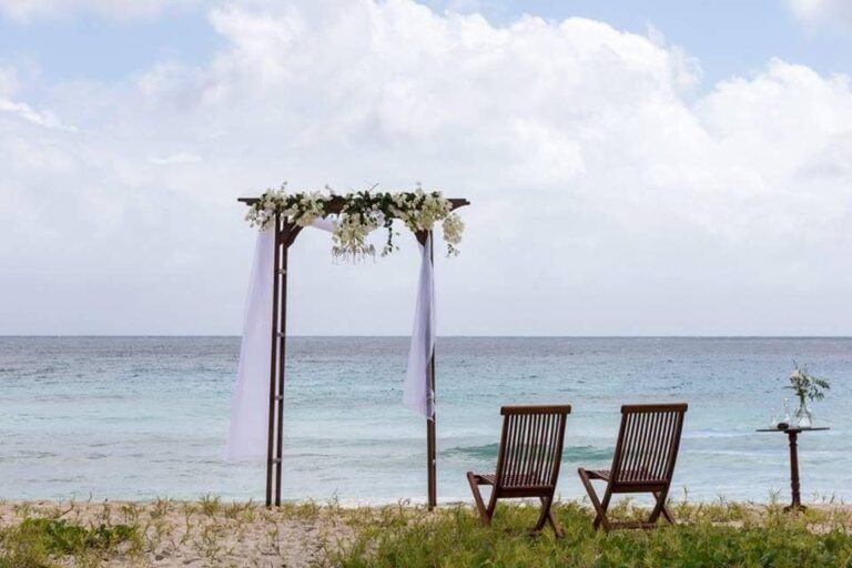 A wooden arch with white drapes and flowers beside two wooden deck chairs facing the sea, Elope Barbados