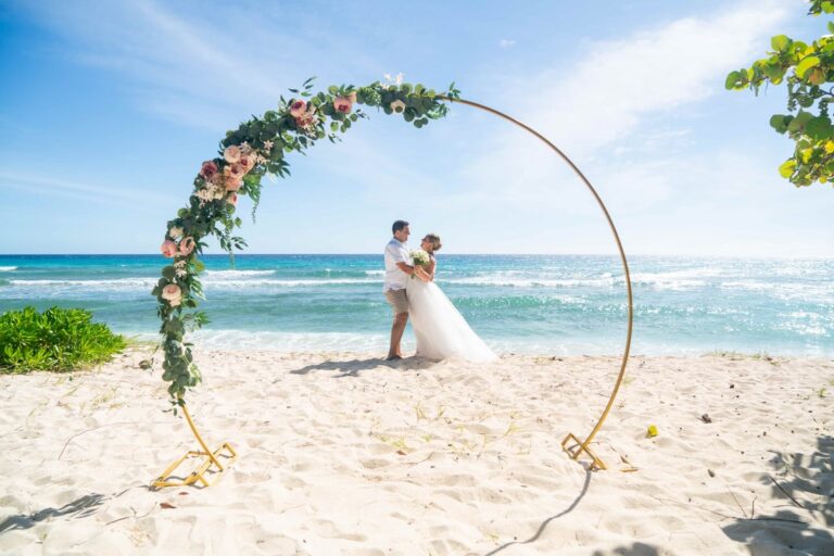 A couple under a round floral arch on a white sand beach with turquoise water beyond, Elope Barbados