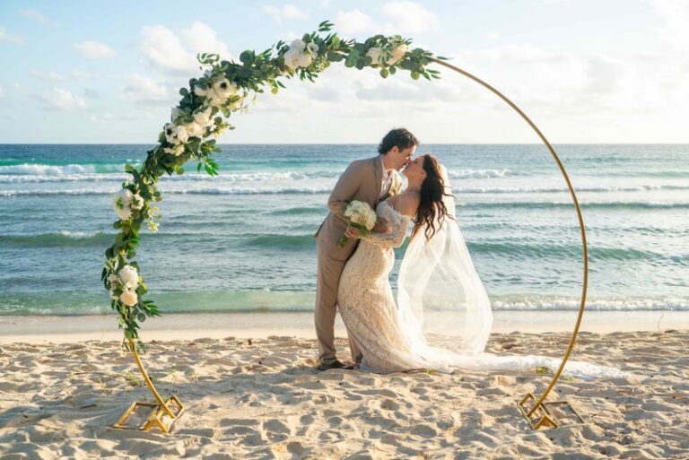 A couple kissing in front of a gold circular arch dressed with roses and greenery on the shore, Elope Barbados