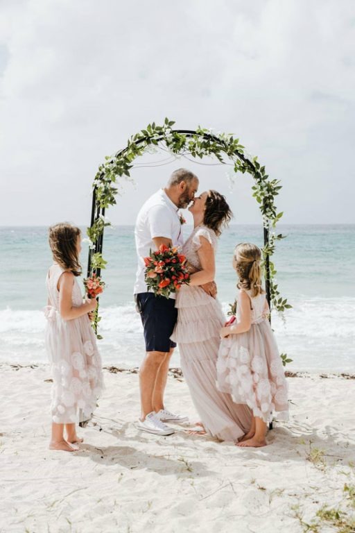 A couple kissing under a green arch on the beach with two young girls in blush dresses beside them, Elope Barbados