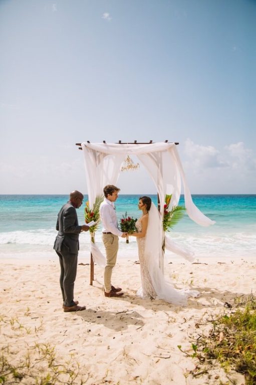 An officiant and couple under a white canopy arch with tropical flowers on the sand, Elope Barbados