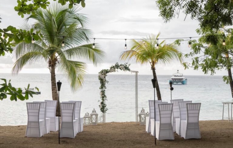 Tables with white chair covers and a flower-dressed arch beneath palms on a beachfront, with festoon lights overhead, Elope Barbados