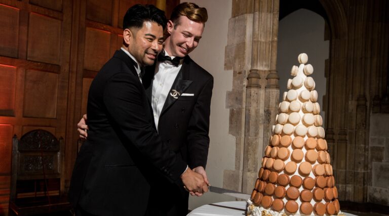 Two grooms in black tie beside a macaron tower, the medieval stone arch of the Great Hall behind, Eltham Palace