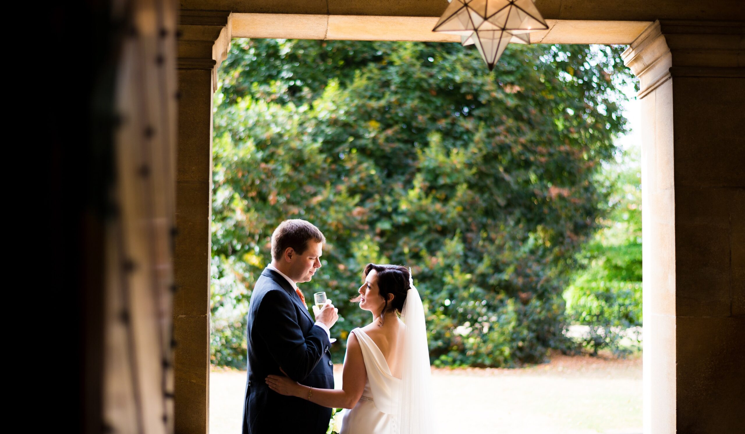 A couple beneath a stone garden loggia, a star lantern above and summer greenery beyond, Eltham Palace