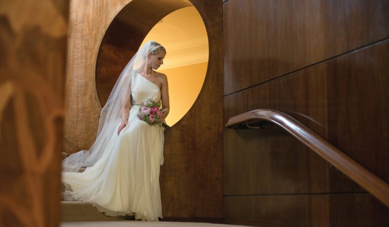 A bride on the curved staircase of the Entrance Hall, beside its round marquetry doorway, Eltham Palace
