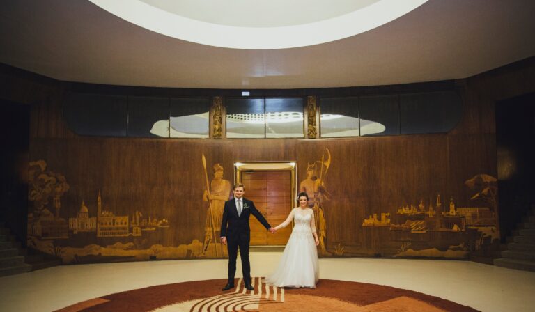 A couple hand in hand under the dome of the Entrance Hall, inlaid cityscapes on the walls, Eltham Palace