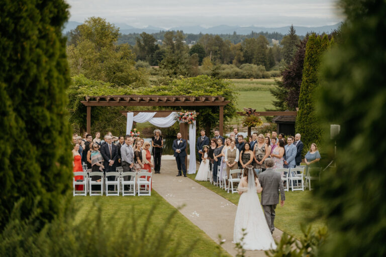 Couple at the end of a long garden aisle beneath a pergola, Emett Joseph Photography