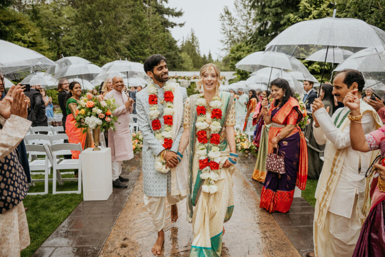 Couple wearing floral garlands walking the aisle between guests under white umbrellas, Emett Joseph Photography