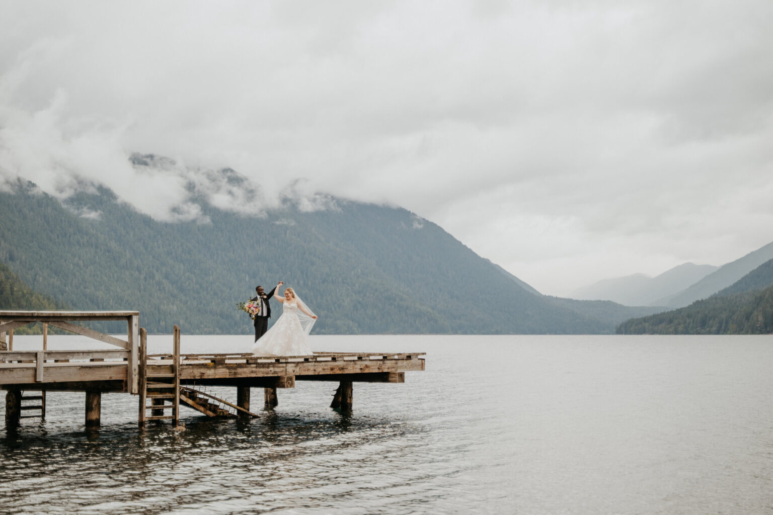 A couple at the end of a lake dock beneath cloud-covered mountains, Lake Crescent