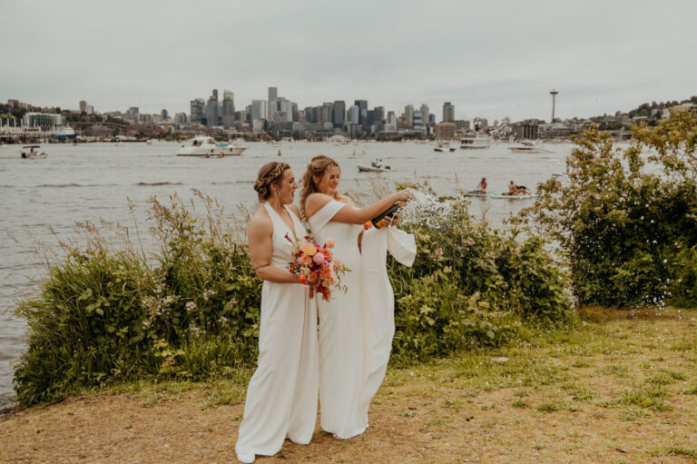 Two brides in white on a waterside lawn with the Seattle skyline beyond, Emett Joseph Photography