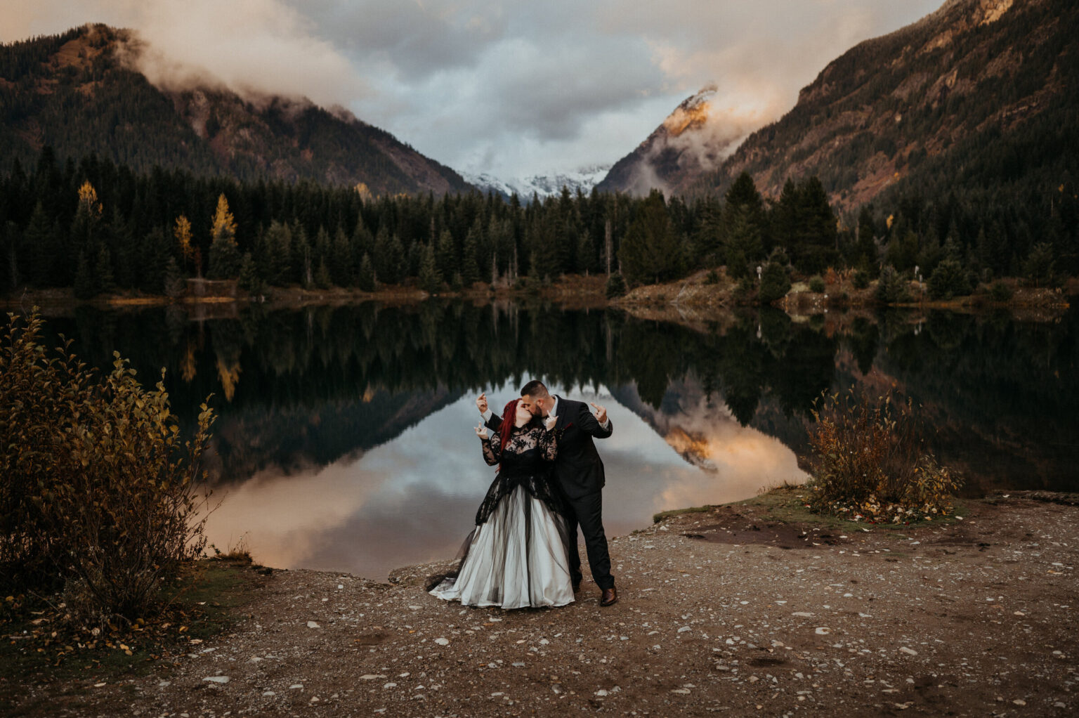 A couple at the edge of a mountain lake at dusk, peaks reflected in the water