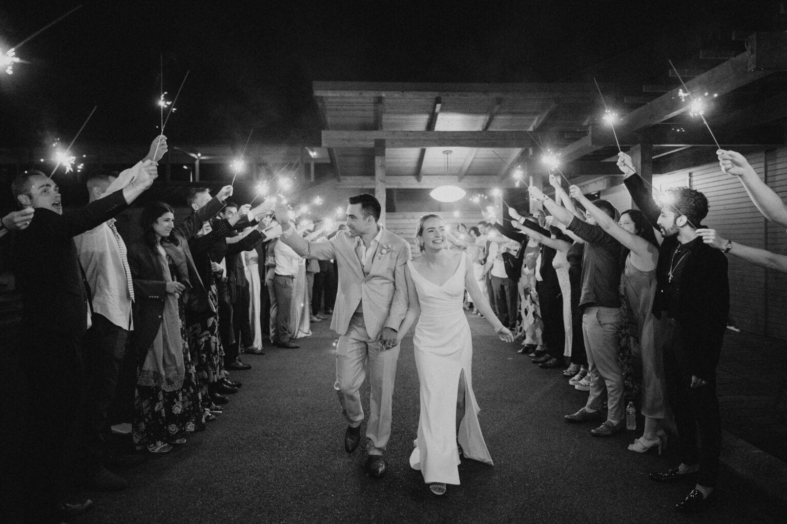 A bride and groom walking between lines of guests holding lights, in black and white