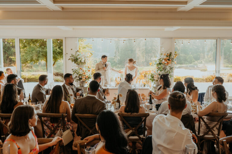 Guests seated for a reception in a bright glass-walled hall beside the lake, Emett Joseph Photography