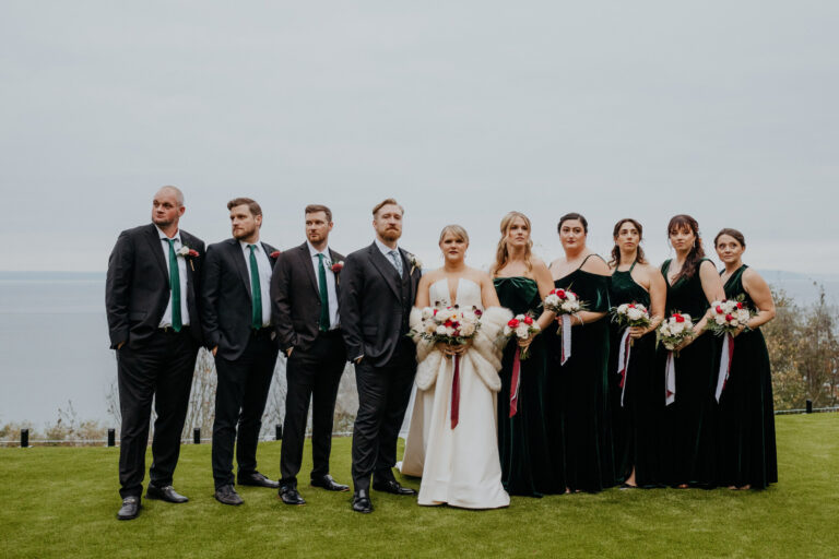 Wedding party in dark suits and black dresses on a lawn by the water, Emett Joseph Photography