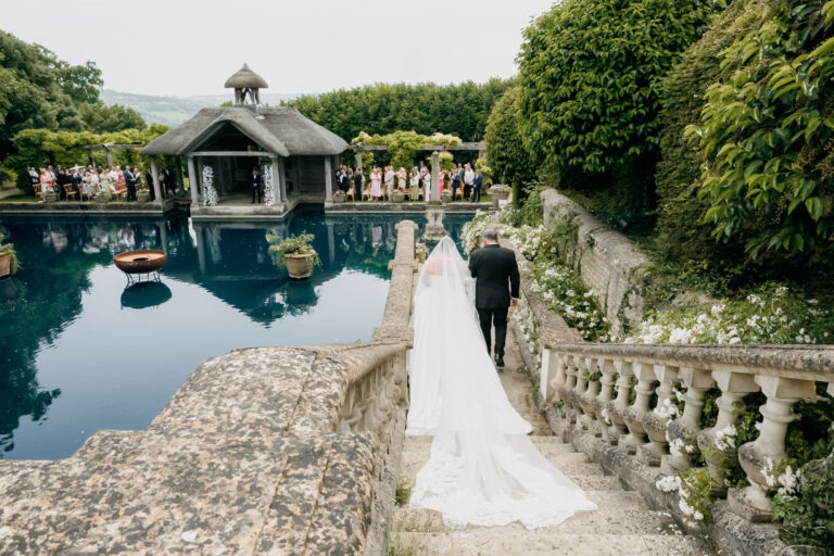 A bride and groom walking the balustraded path beside the water garden at Euridge Manor, guests gathered at the pavilion, Emma-Jane Photography