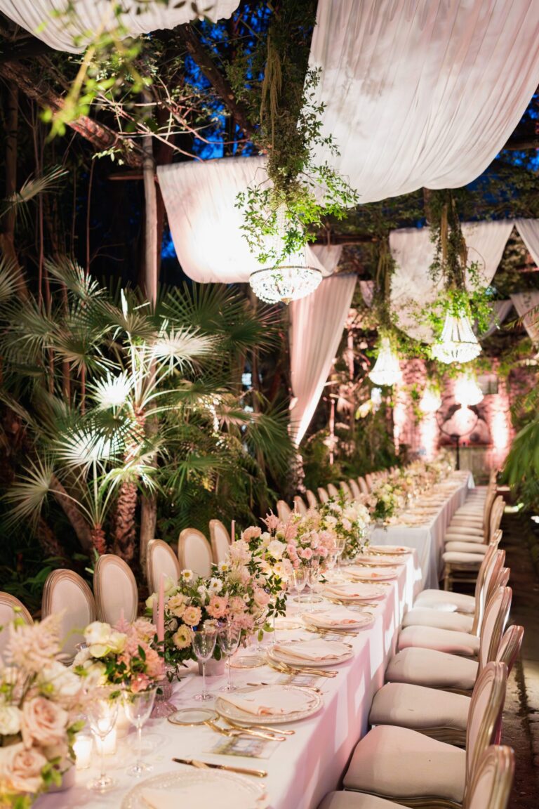 Pink drapery and chandeliers over the long table, palms behind, Ester Chianelli Weddings & Events