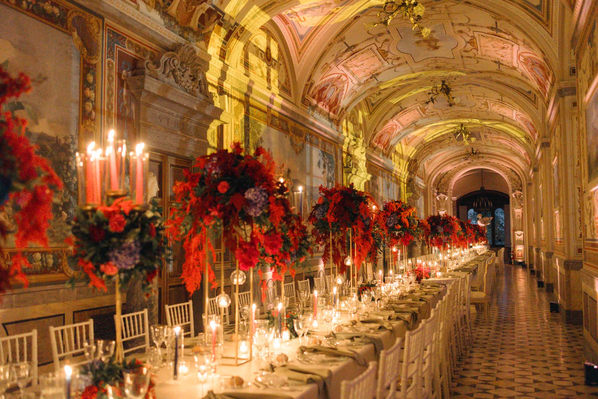 A long candlelit table under the frescoed gallery at Castello di Torre Alfina, with tall red and coral floral arrangements, Eventilab Weddings