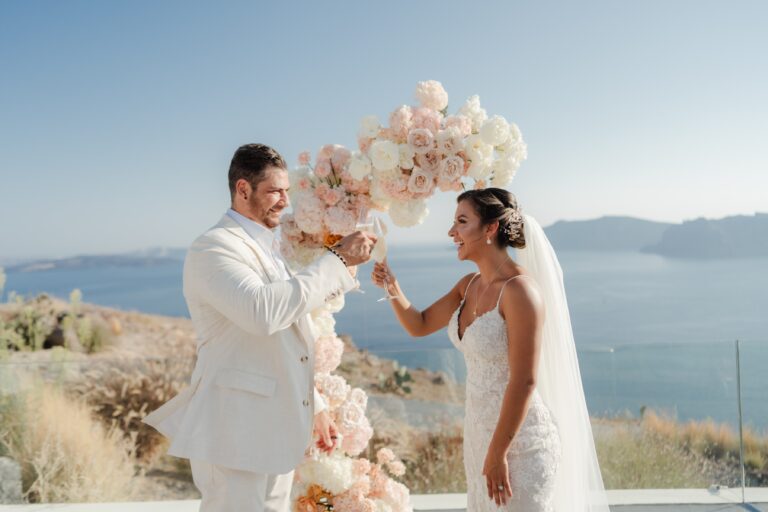 A couple at a clifftop elopement ceremony beside an arch of pale roses, with the caldera behind, Eventuries Wedding Planning and Styling