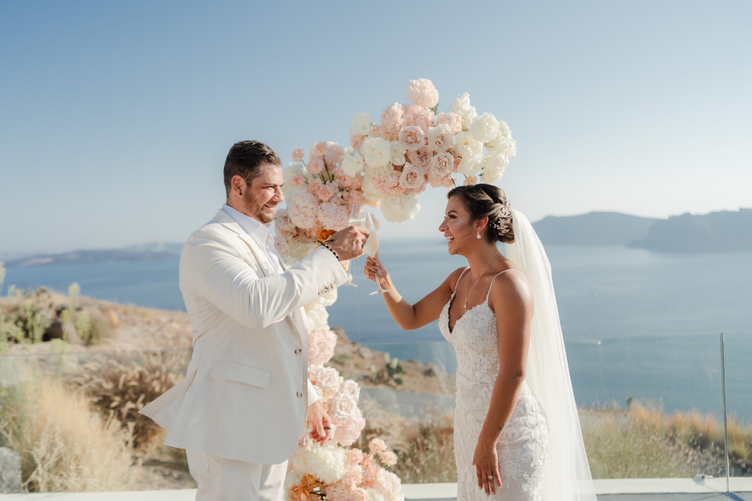 A couple at a clifftop elopement ceremony beside an arch of pale roses, with the caldera behind, Eventuries Wedding Planning and Styling